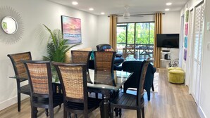 A dining area with dark wooden chairs and a glass table, a television on a stand, and a large window with curtains. - Minutes to Sanibel with Heated Pool, Tennis & Beach Gear (Fort Myers)