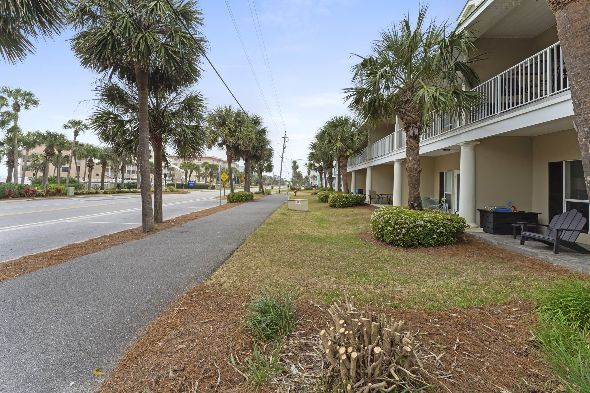 A hotel with a covered outdoor seating area, palm trees, and a sidewalk.