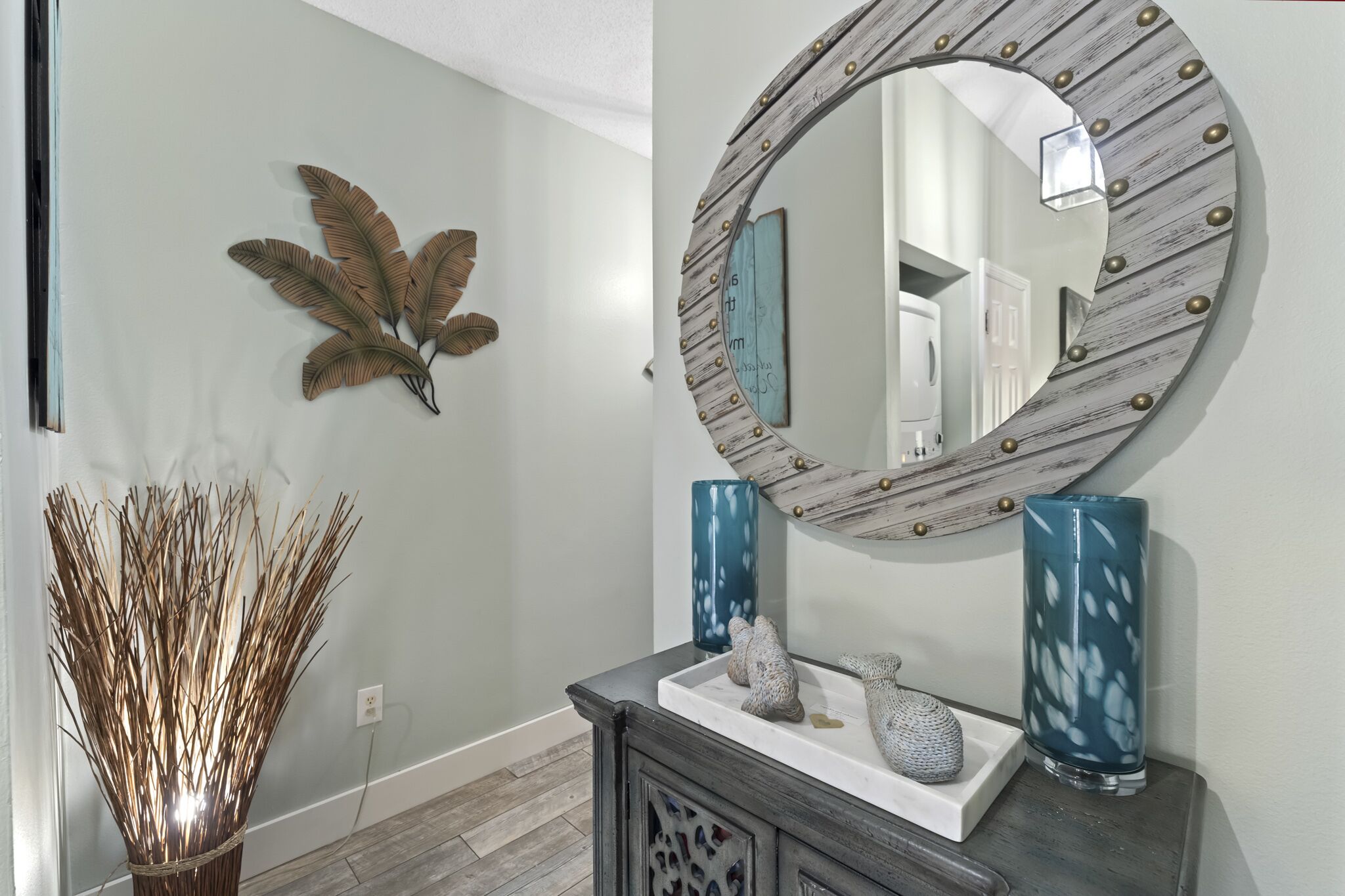 Un couloir agrémenté d’un élément décoratif mural, d’un miroir au cadre rustique et d’un vase posé sur une console.