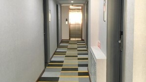 Ein Hotelgang mit Schachbrettmuster auf dem Boden, Türen an beiden Seiten und einem Lüftungsgitter an der rechten Wand.