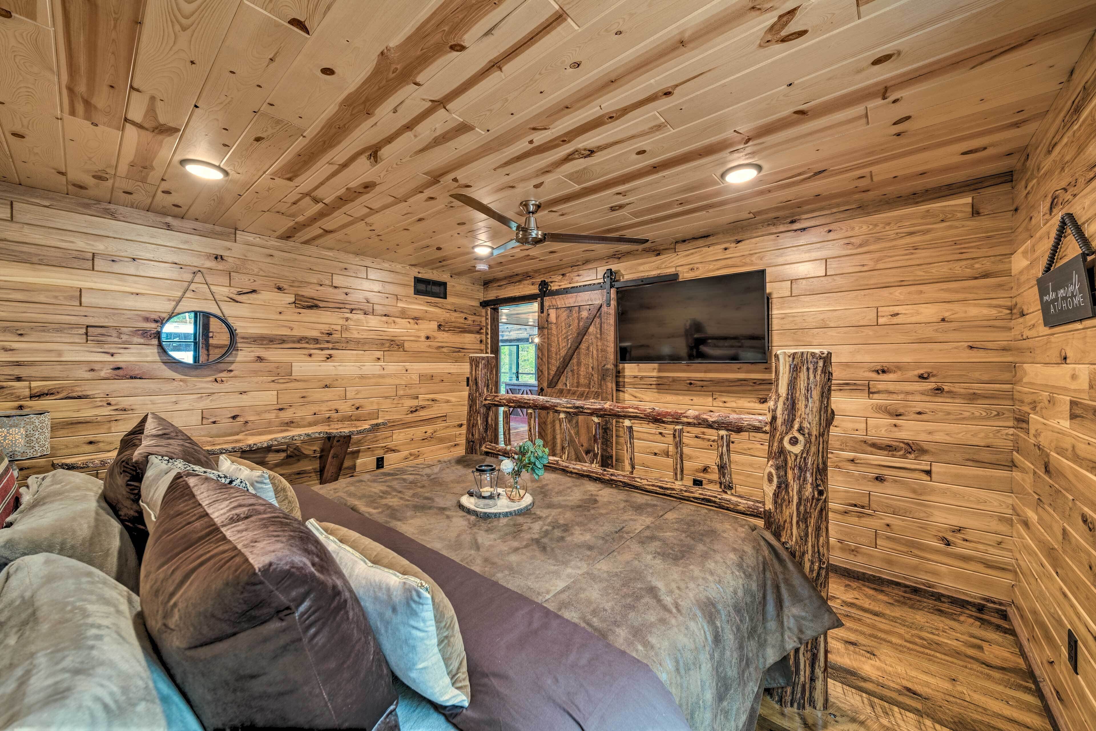 L’intérieur d’un chalet accueillant, avec des murs en bois, un ventilateur de plafond, un grand lit avec des oreillers, une petite table avec une bougie et un téléviseur fixé au mur.