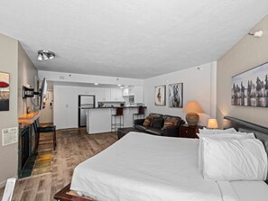 Ein modernes Hotelzimmer mit einem großen Bett, einer kleinen Küchenzeile, einem Essbereich und einer Wohnlounge inklusive Sofa und Fernseher.