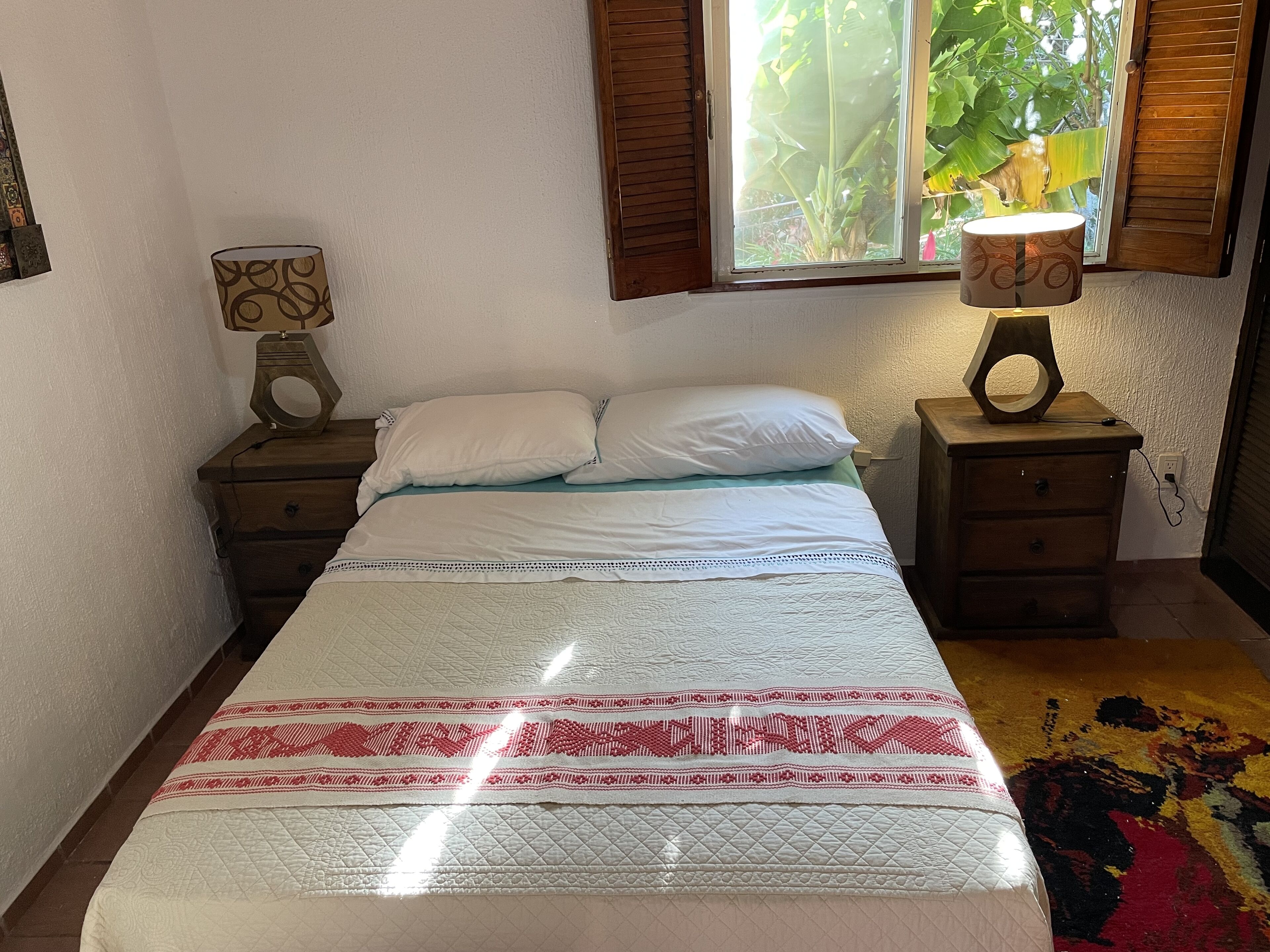 A bedroom with a bed, two bedside tables, a window with blinds, and a lamp on one of the tables.
