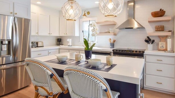 A modern kitchen with white cabinetry, a stainless steel refrigerator, a gas stove, and two wicker chairs. - Steps to Oyster Pond Private Dock! (Chatham)