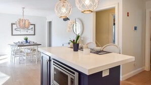 A modern kitchen with a white island, pendant lights, and a dining area with white chairs. - Steps to Oyster Pond Private Dock! (Chatham)