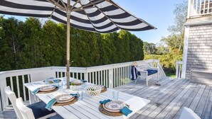 A deck with a white table set for dining, surrounded by white wicker chairs, and a striped umbrella providing shade. - Steps to Oyster Pond Private Dock! (Chatham)