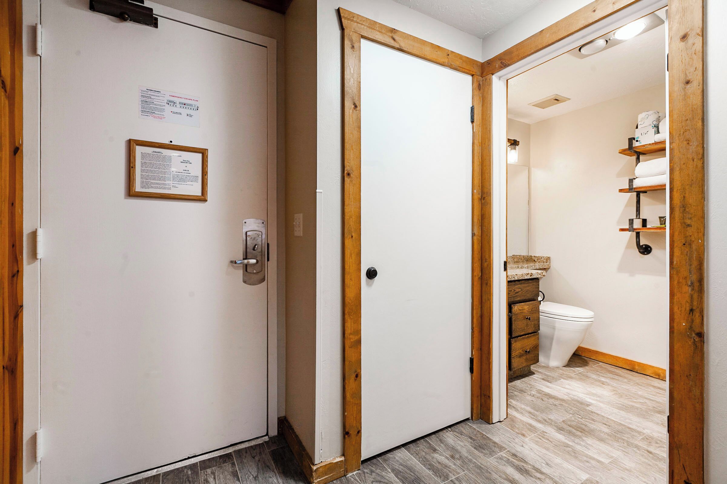 Een kamer met een houten deur, een witte muur met een bericht en een badkamer met toilet en planken.