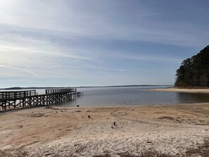 Una playa con un muelle de madera que se adentra en aguas tranquilas, rodeada de arena y vegetación dispersa.