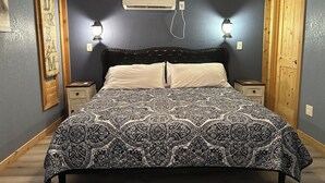 Une chambre avec une couvre-lit à motifs, une tête de lit noire, deux lampes de chevet, un appareil de climatisation et des murs en bois.