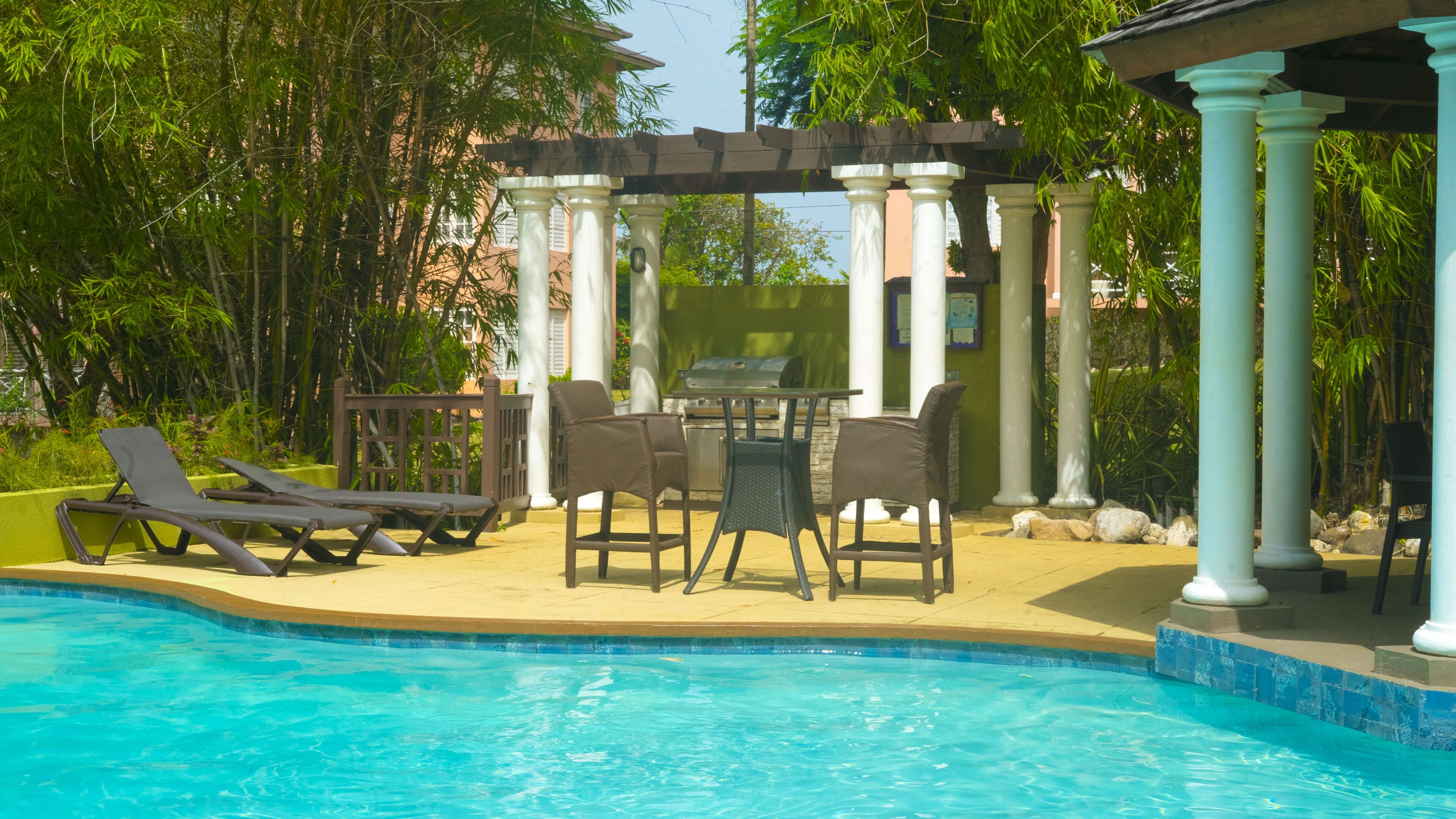 A pool area with lounge chairs, a barbecue grill, and a pergola with columns.
