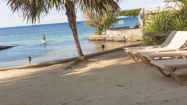 A beach with clear blue water, a person surfing, and lounge chairs along the shore. - Penthouse villa on the beach, chef services available- extra cost.  (Ocho Rios)
