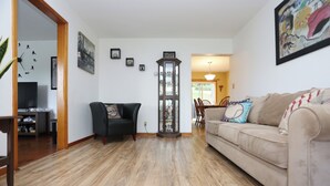 Una sala de estar con un sofá color beige, un sillón negro y piso de madera. En la pared, hay un reloj de madera alto y oscuro.