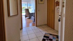 Un pasillo con suelo de baldosas, una puerta de madera, un cuadro en la pared y un tapete que lleva a una habitación con una silla y una ventana.