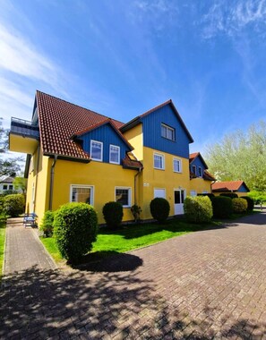 A two-story yellow and blue house with a red-tiled roof, a paved driveway, and well-maintained hedges.