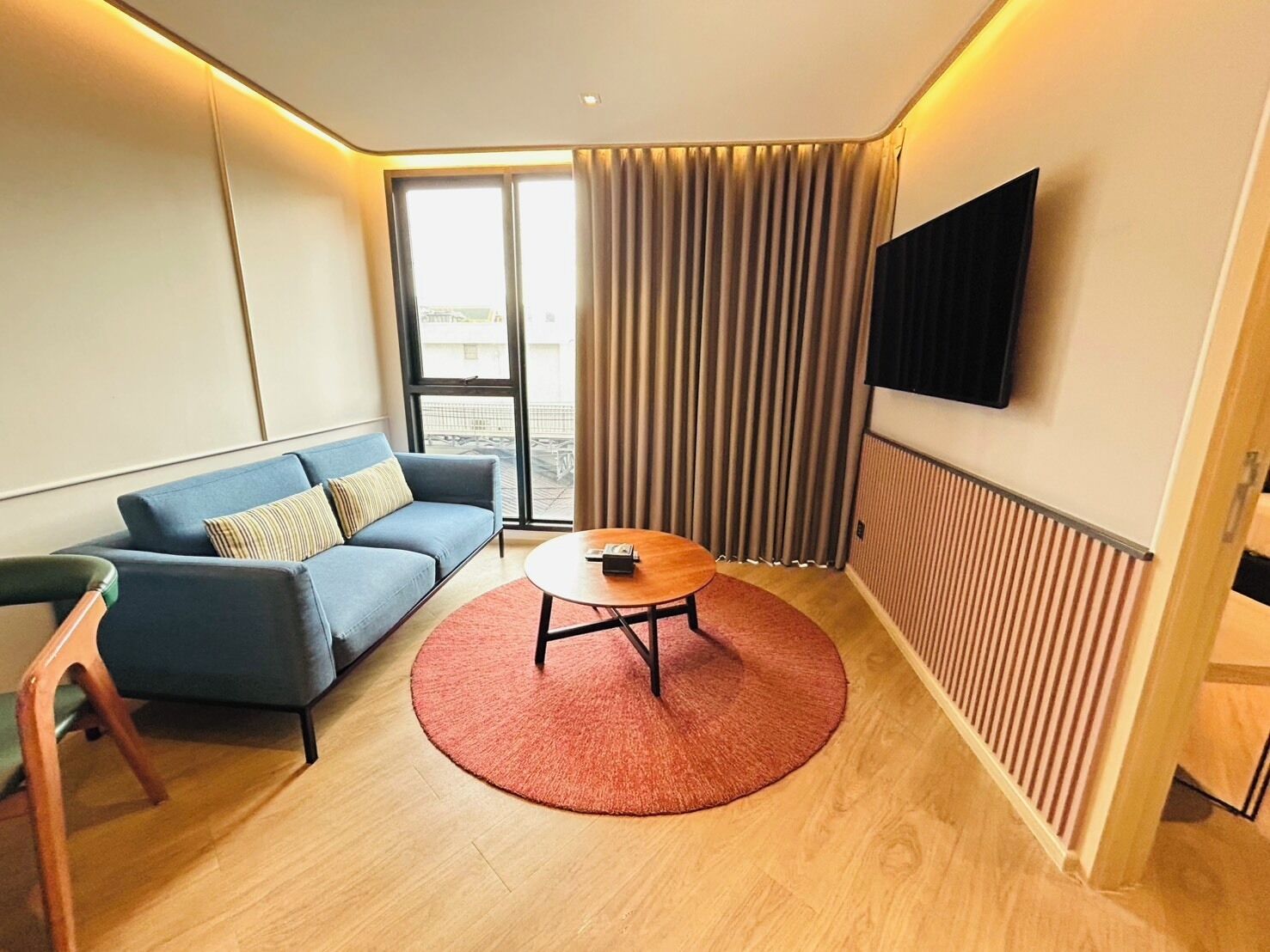 Una habitación de hotel moderna con un sofá azul, una mesa redonda de centro, un televisor montado en la pared y un amplio ventanal con cortinas.