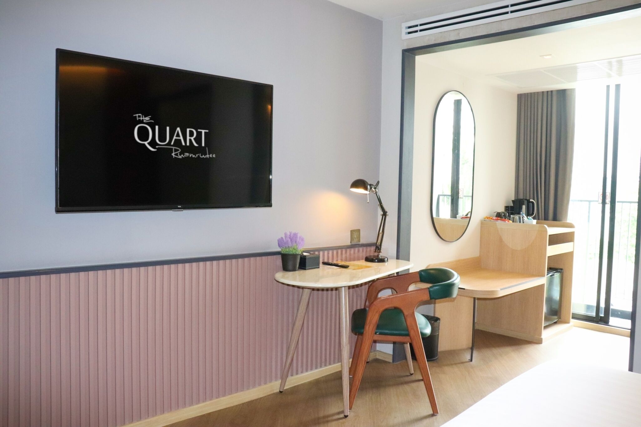 Habitación de hotel con una televisión mostrando "The QUART Reimagine", un escritorio con una lámpara, una silla, una pequeña planta en maceta y un espejo grande.