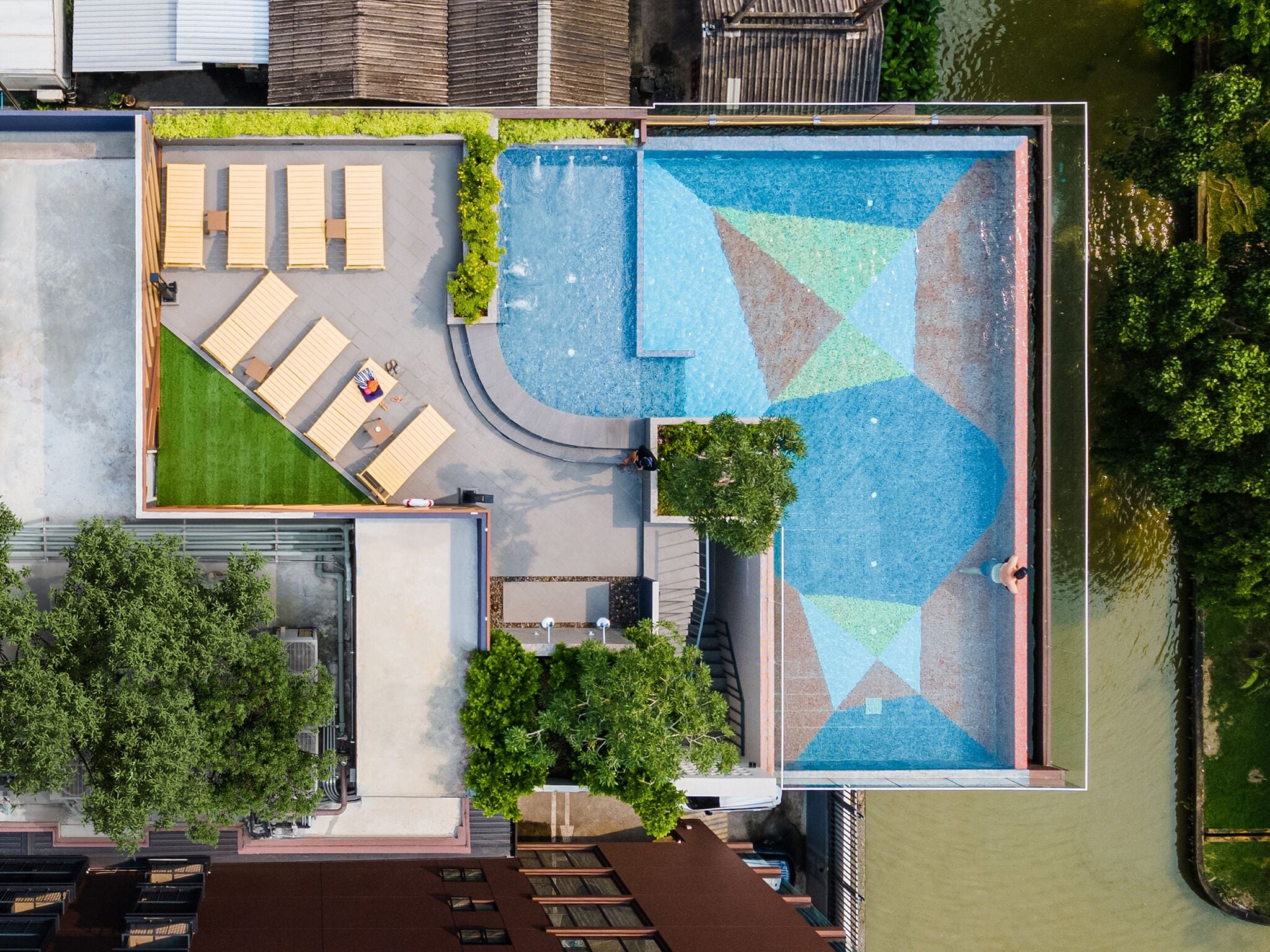 An aerial view of a rooftop pool area with a geometric tile design, seating, and greenery.