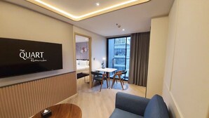 Een moderne hotelkamer met een flatscreen televisie, een bank, een eettafel met stoelen en een uitzicht op de stad door grote ramen.