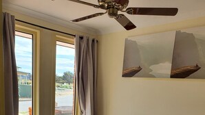 A bedroom with a bed, wooden headboard, nightstands, a ceiling fan, and a view of the outdoors through large windows. - Stylish house in the middle of the Links Lady Bay Golf Course (Normanville)