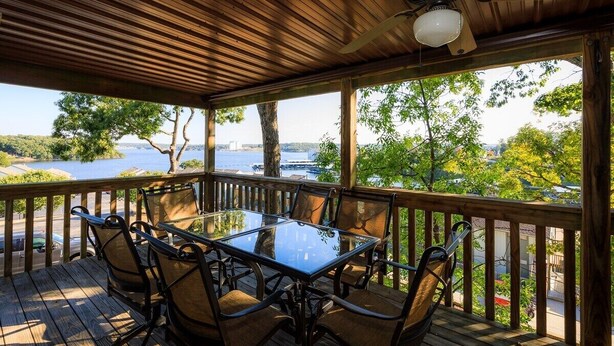A covered outdoor deck with a glass table and chairs, overlooking a body of water and greenery.