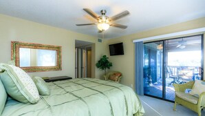 A bedroom with a large bed, a ceiling fan, a TV, and a sliding glass door leading to an outdoor area with furniture.