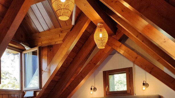 A cozy attic bedroom with a wooden ceiling, a large bed with white bedding, two wall-mounted light fixtures, and a window with a view of the outdoors. - Bamboo I - tucasaenlospirineos (Escarrilla)