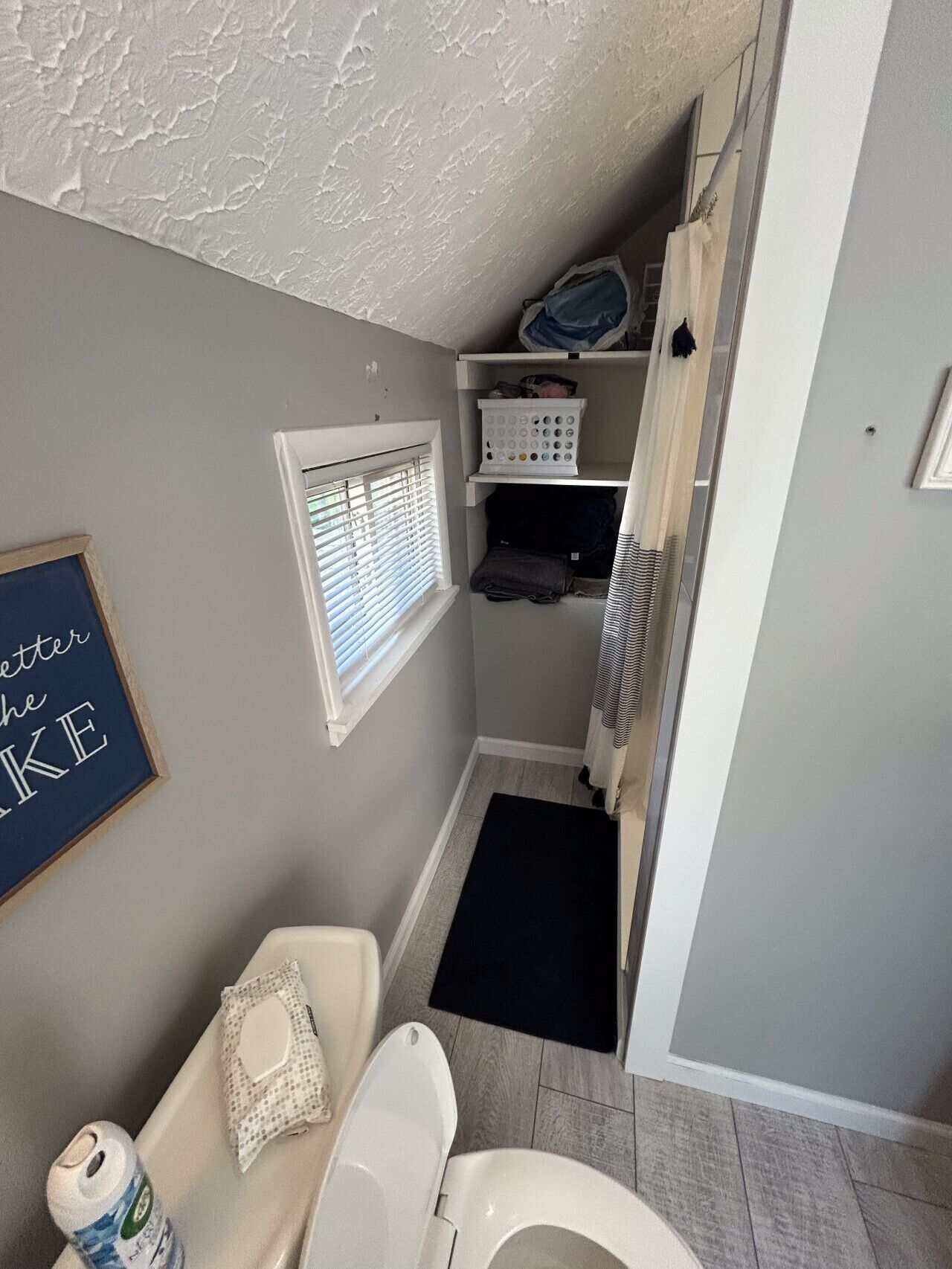 A compact bathroom with a toilet, a small window, a shelf with storage, and a shower area.