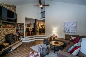 A cozy living room with a stone fireplace, a television, a dining area, and a kitchen visible through an open door.