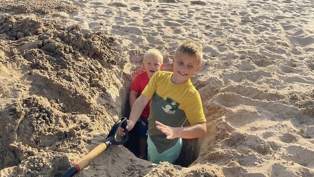 Zwei Kinder spielen in einer Sandkiste am Strand, mit blauem Himmel und einem entfernten Leuchtturm.