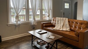 Ein Wohnzimmer mit braunem Ledersofa, einem Holztisch und einem Fenster mit durchsichtigen Vorhängen.
