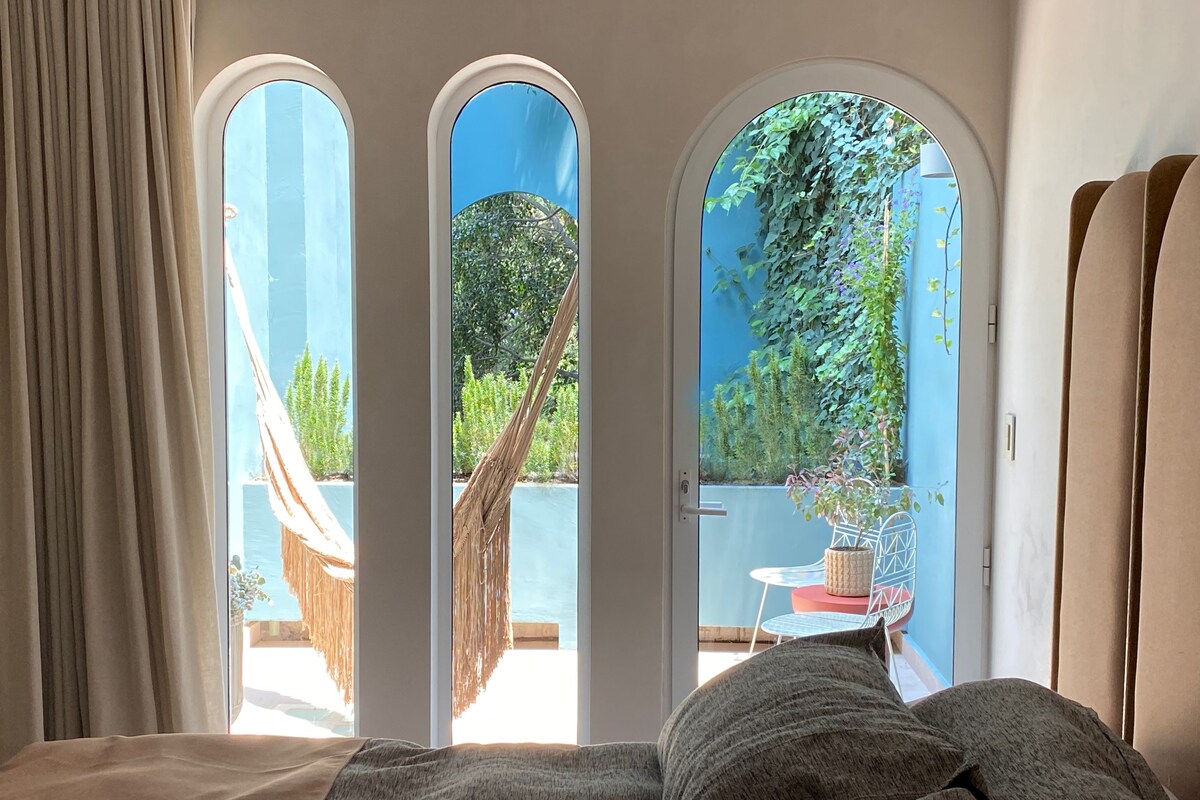 A room with a bed, a view through arched windows to an outdoor area with a hammock, a table, and potted plants.