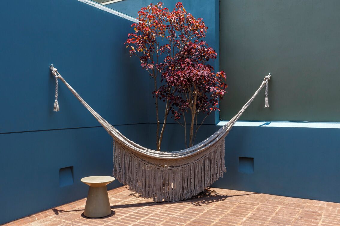 Una hamaca con flecos, una pequeña mesa redonda y un árbol de hojas rojas frente a un muro azul.