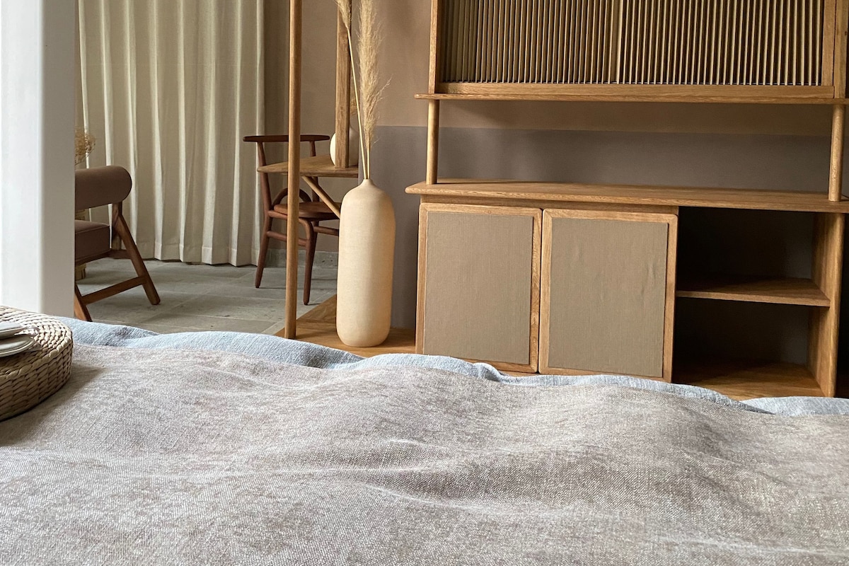A room with a bed, a wooden desk, a chair, and a large vase with dried plants.