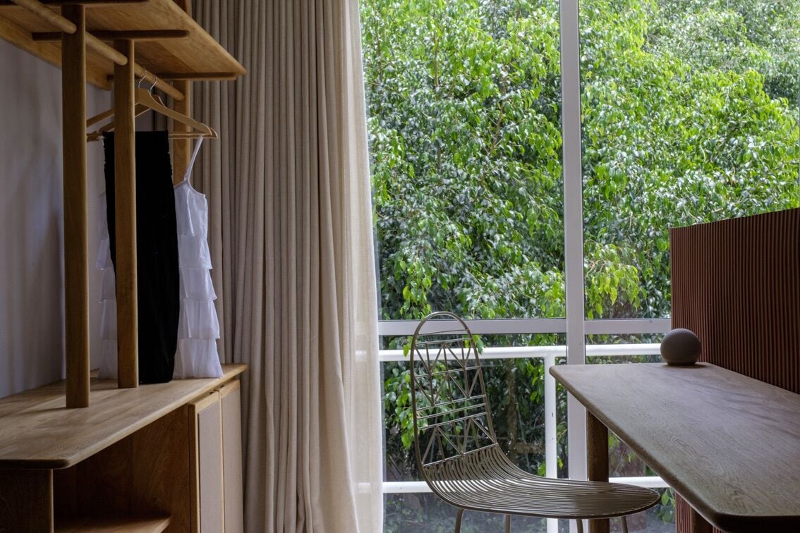 Una habitación con una mesa de madera, una silla metálica, un estante de madera con ropa, y vistas de un follaje verde exuberante a través de un ventanal grande.