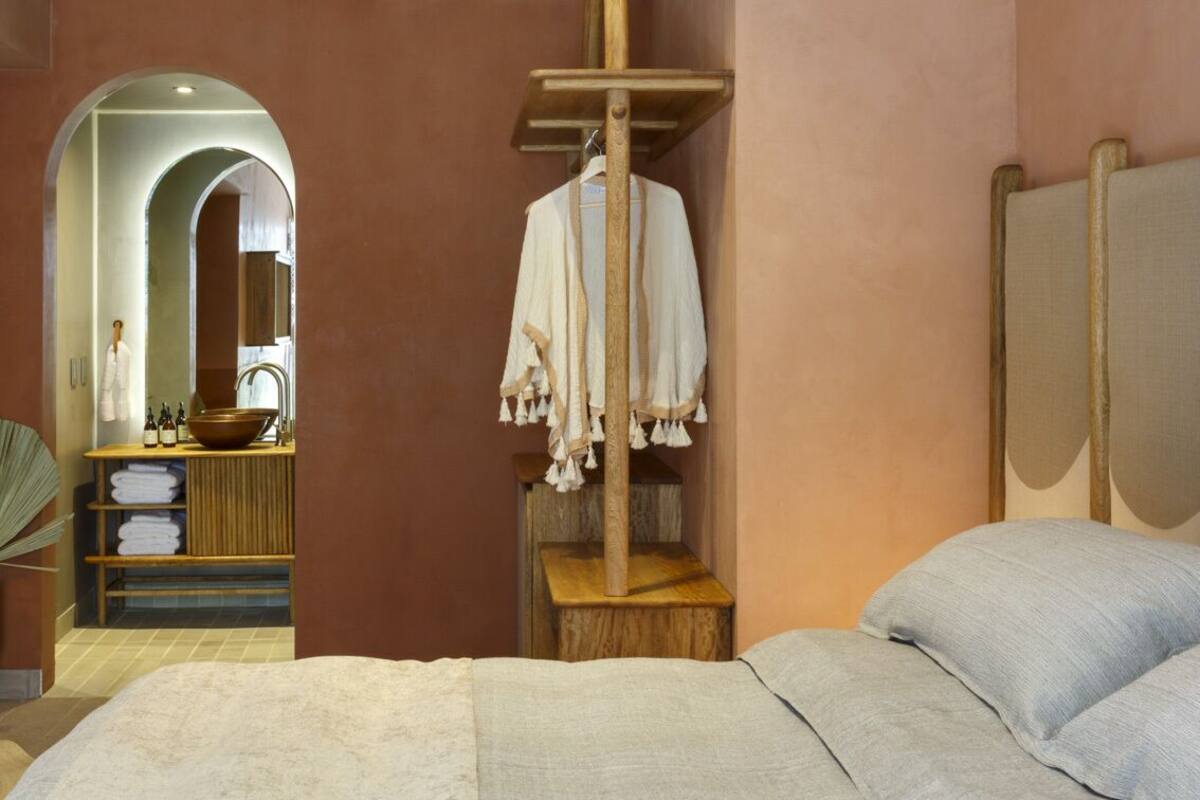 A bedroom with a bed, a wooden shelf with a hanging robe, a wooden stool, and a mirror.