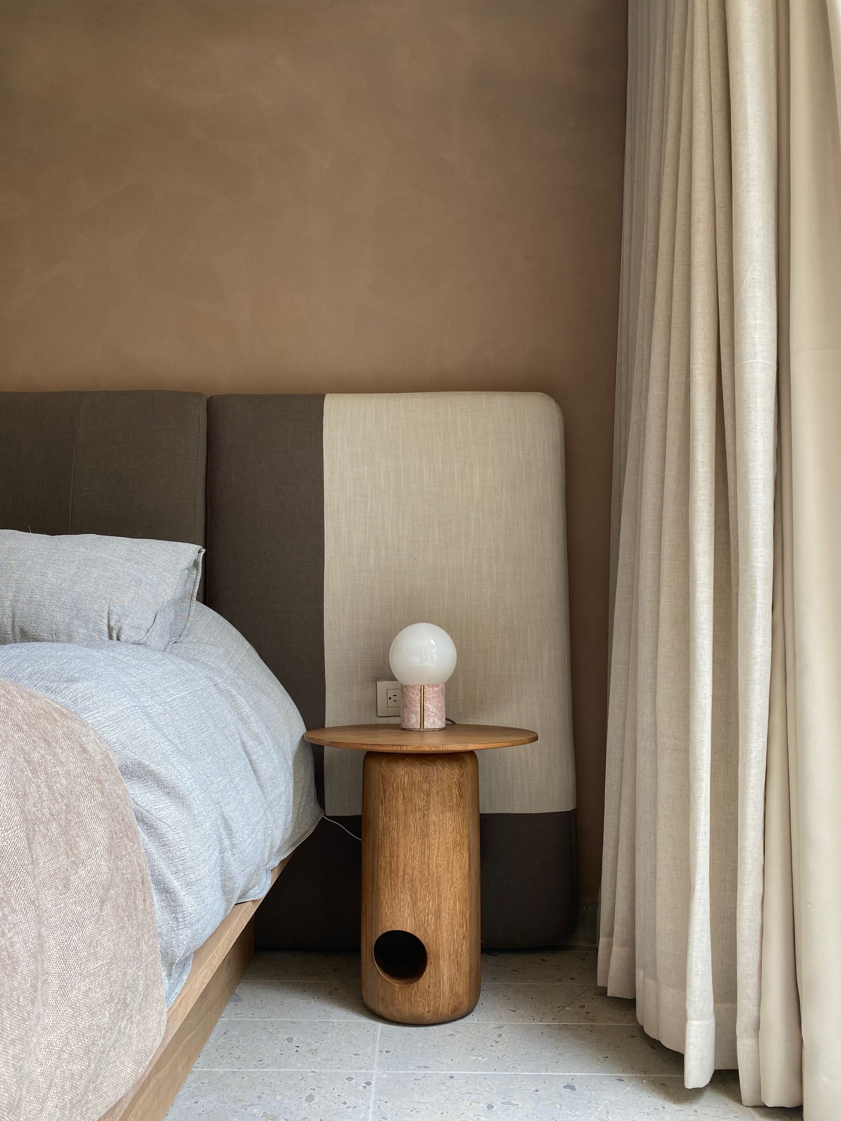 A bedroom with a bed, a round wooden nightstand with a lamp, a wall-mounted shelf, and a window with curtains.