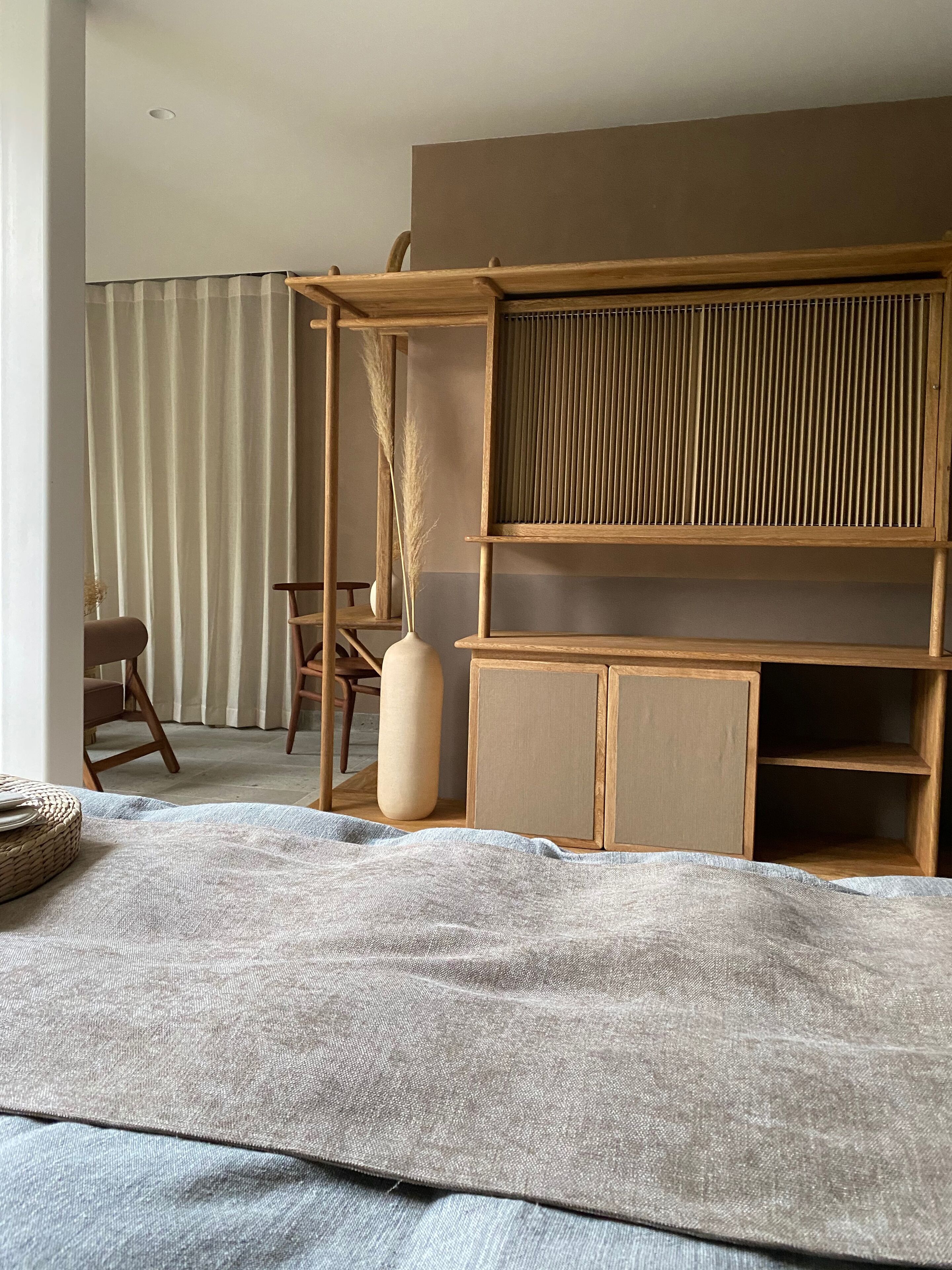 A room with a bed, a wooden desk, a chair, and a large vase with dried plants.