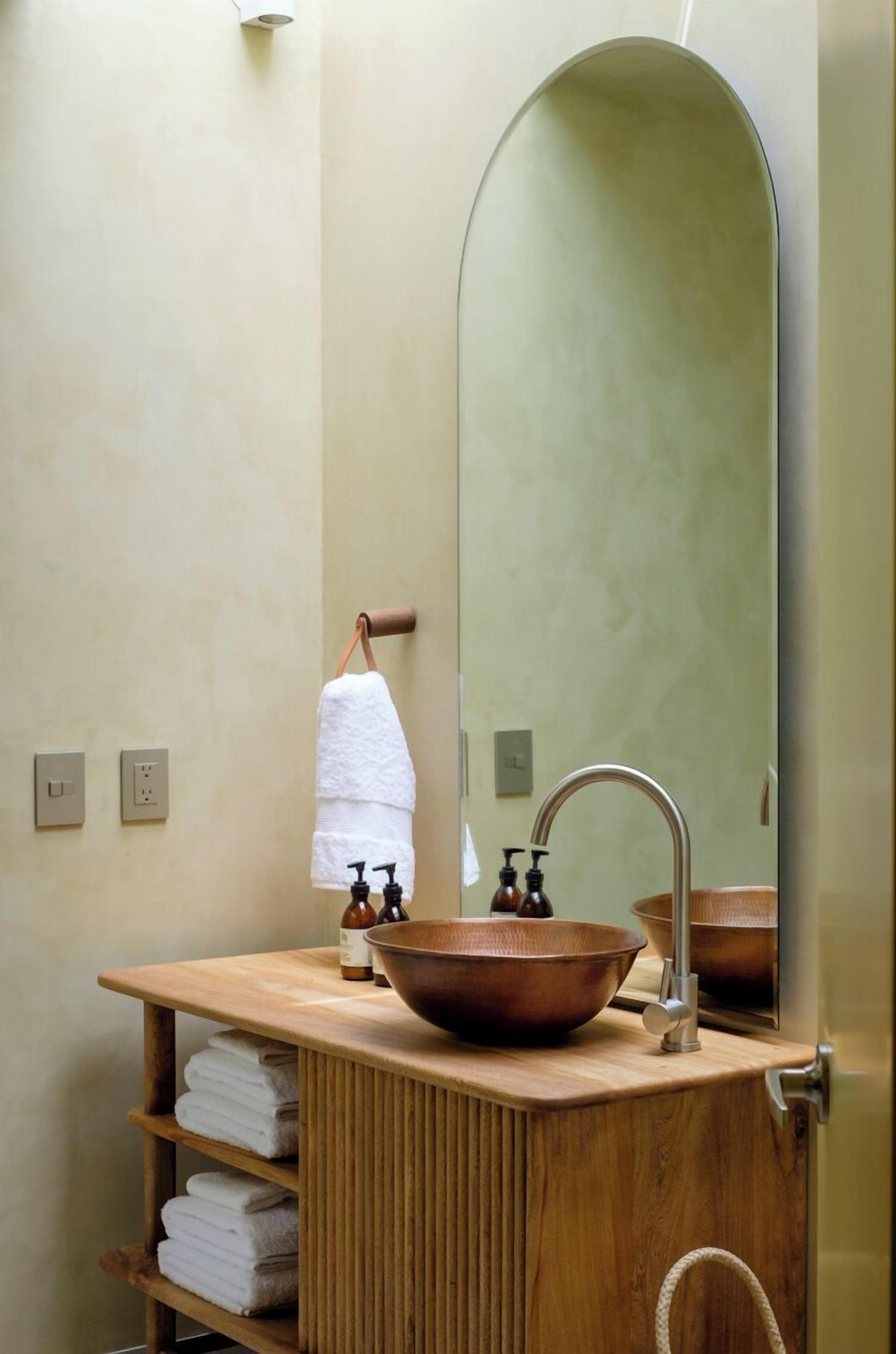 Un baño con tocador de madera, lavamanos de cobre, un espejo grande y un perchero para toallas.