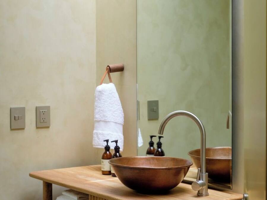 Un baño con tocador de madera, lavamanos de cobre, un espejo grande y un perchero para toallas.