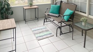 Ein Wintergarten mit großem Fenster, Deckenventilator, Sessel mit Kissen, einem kleinen Tisch und einer Topfpflanze.