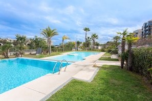 A rectangular swimming pool with a metal ladder, surrounded by grass and palm trees, with buildings in the background.