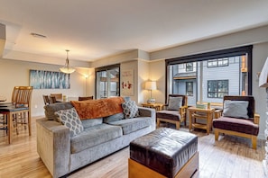 Amplia sala de estar con un sofá gris, un reposapiés de cuero, muebles de madera y ventanas grandes.