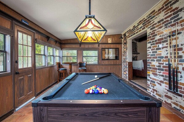 A pool table with balls, a hanging light fixture, and a brick wall. - Madeira Haus | Pool & Hot Tub | Pet-friendly (Fredericksburg)