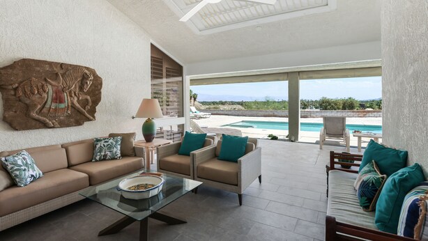 A spacious living area with a sofa, armchairs, and a coffee table. A large artwork is on the wall, and there is a view of a pool and landscape outside. - Casual Elegance in the Desert | A Louisa Davis Property (Rancho Mirage)