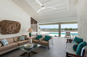 A spacious living area with a sofa, armchairs, and a coffee table. A large artwork is on the wall, and there is a view of a pool and landscape outside. - Casual Elegance in the Desert | A Louisa Davis Property (Rancho Mirage)