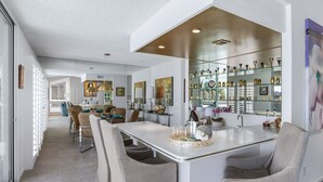 A modern kitchen with a central island, dining area with chairs, and a glass-fronted display cabinet with decorative items. - Casual Elegance in the Desert | A Louisa Davis Property (Rancho Mirage)