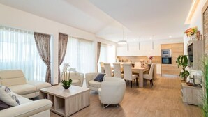 Un salon moderne comprenant un canapé, un fauteuil et une table basse, un coin repas avec des chaises, et une cuisine équipée d’appareils électroménagers.