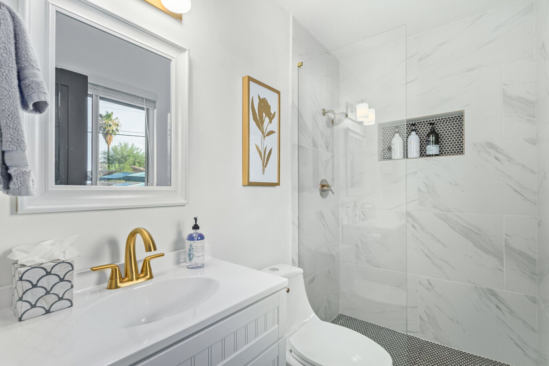 Un bagno con lavandino bianco, rubinetto dorato e water. C'è un quadro appeso alla parete e una finestra con vista su palme.