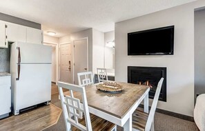 A modern kitchen with white cabinets, a built-in refrigerator, a dining table with chairs, a flat-screen TV mounted above the fireplace, and a small decorative tray on the table.