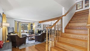 Amplia sala de estar con escalera de madera, muebles modernos y ventanas grandes.